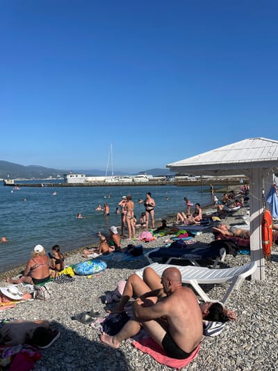 The image depicts a crowded pebble beach on a sunny day. The water is calm and blue, with several people swimming and wading. Beachgoers are spread across the shore, some reclining on beach chairs or towels, while others are in the process of changing or interacting. A white canopy extends over a section of the beach, providing shade. A life preserver hangs from the canopy structure.

In the foreground, a bald man wearing black swim trunks sits on a pink towel, leaning forward. Next to him, another person with dark hair tied up and wearing glasses is seated on a white beach chair. Further down the beach, a woman in a patterned bikini is standing, while other individuals are lying down or sitting.

The background shows a clear blue sky and mountains in the distance. A structure that appears to be a pier or a seawall stretches along the coastline, with a sailboat docked beside it. The presence of beach umbrellas, towels, and swimwear indicates a typical summer leisure activity. The bright sunlight suggests it is midday. The scene is lively, with many people enjoying the beach and the water.