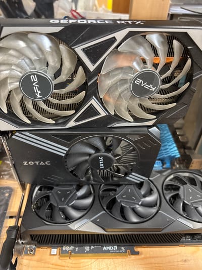 The image shows three computer graphics cards stacked vertically. The top card has "GEFORCE RTX" printed on it and features two fans with circular black emblems that read "EVGA". The middle card, branded "ZOTAC", also has two fans, and the bottom card has "AMD" printed on its circuit board. The graphics cards are placed on a wooden surface, possibly a workbench, in what appears to be an indoor setting. The background is out of focus, but it suggests an environment with other electronic components or equipment. There are no people visible in the image. The image does not provide any information about the location being Mülheim an der Ruhr, Germany.