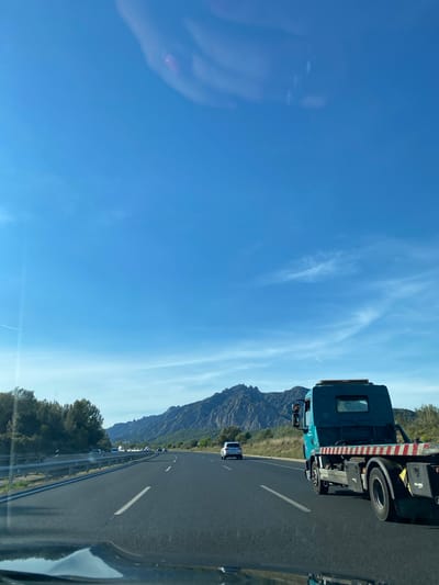 A slightly tilted, broad shot captures a clear blue sky over a highway, reflecting in the car window on the left. The highway stretches into the distance, lined with guardrails and scattered vegetation on one side. A tow truck is positioned on the right of the frame, followed by a mountain range in the background. The location appears to be along a road near Esparreguera, Spain, suggested by the mountainous terrain in the distance.