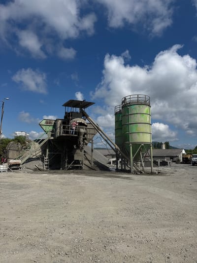 The image shows a concrete mixing plant and two tall green silos at an industrial site. A person is visible on an elevated platform of the mixing plant, appearing to be working. There are piles of gravel and construction materials around the machinery. In the background, there are several low-rise industrial buildings with grey roofs, and a truck is parked to the right. Hills and trees are visible in the distance. The sky is blue with scattered white clouds. The ground is rough and covered with gravel and dirt. This appears to be an outdoor industrial facility, possibly located in Zugdidi, Georgia, given the context.