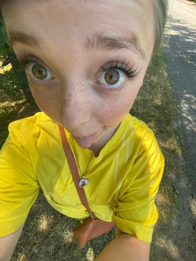 This is a close-up, high-angle, selfie-style shot of a young woman in Nova Praha, Ukraine. The woman, who appears to be a teenager or young adult, is wearing a bright yellow t-shirt with thin white stripes and a dark brown crossbody bag with a gold buckle. She has a pendant necklace with a circular charm featuring a picture of a horse. Her left eye, adorned with long eyelashes, is prominently visible, and her tongue is sticking out slightly. She has light skin with freckles across her nose and cheeks. The background suggests an outdoor setting with dry grass and shadows cast by trees, indicating it is daytime, likely a sunny day. The orientation of the image is such that the woman's head is at the top left of the frame. No visible text is present in the image.