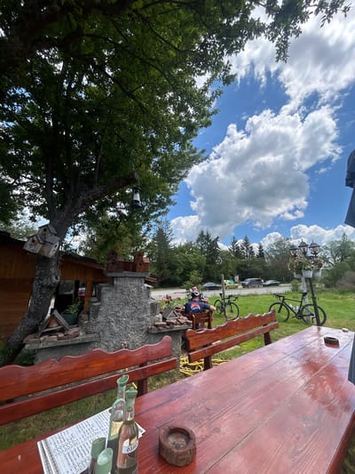 The image shows an outdoor scene, potentially in Meshtitsa, Bulgaria, though this cannot be definitively confirmed from the image alone. A large wooden table and benches dominate the right side of the frame. On the table, there are bottles, and what appears to be a menu or printed paper. Next to the table, there's a stone structure and stacked wood. A cyclist is seen sitting on one of the benches, further away, near a road with parked cars and a streetlight with decorative elements and a bicycle mounted on it. A tree with multiple birdhouses attached to its trunk is visible on the left side. The sky is blue with scattered clouds.