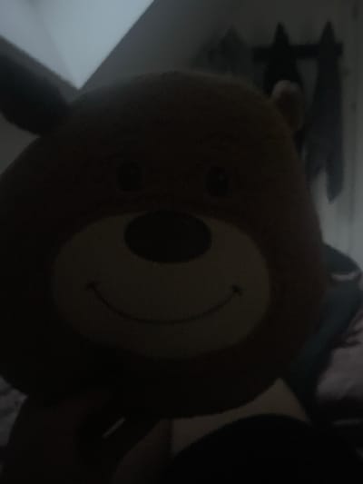 The image is a dimly lit, close-up, and slightly blurry photograph of a brown stuffed bear's head. The bear appears to be very large and soft. Its features, including its eyes, nose, and a wide smile, are visible, but the overall image is dominated by shadows. The background is out of focus, with what appears to be a gray ceiling or wall and some dark, vertical shapes that might be curtains or clothing hanging. The image quality suggests it was taken in low light conditions with a mobile device. The specific location in Paczkowo, Poland, is not discernible from the visual information provided in the image itself.