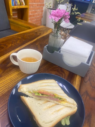 This is a medium-shot, eye-level photograph of a light brown wooden table in a cafe. The table is set with a meal: a dark blue plate holding a halved grilled sandwich, a white ceramic mug filled with a light brown liquid, a grey napkin holder with a stack of white napkins, and a small glass vase containing a single pink flower with thin stems and small white buds.
The sandwich is made with white bread that has grill marks. It is filled with lettuce, tomato, ham, and melted cheese. The mug is simple and fluted in design, with a handle. The napkin holder is rectangular with curved indentations on the sides. The flower is a carnation, with its petals tightly packed and a soft pink hue.
In the background, a bookshelf filled with books is visible, as is a brick wall. A portion of a dark grey upholstered chair is also present. The lighting is soft, suggesting an indoor setting during the day. There are no people visible in the frame.
The image does not provide any specific information about the location being in Kolomna, Russia.