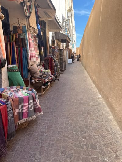 The image depicts a narrow, cobblestone alleyway in Essaouira, Morocco, lined with shops on the left. The shops display a variety of textiles, including colorful scarves and blankets, hung on racks and folded in baskets. The buildings have traditional Moroccan architecture, with textured walls and arched doorways. Sunlight casts shadows on the alley, highlighting the contrast between light and dark. In the distance, a few people are visible walking through the alley. The right side of the alley is dominated by a tall, smooth, sandy-colored wall, casting a long shadow over the cobblestones. The sky above is clear and blue with a few wispy clouds. The overall impression is one of a bustling marketplace in an ancient city.