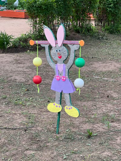 The image shows a wooden cutout of a gray bunny in a park. The bunny is wearing a purple dress with pink straps and yellow shoes. It is holding a bar across its chest, from which hang four colorful balls: yellow, red, green, and white. The bunny has pink ears, blue eyes, a red mouth, and whiskers. The background consists of dirt, grass, and green bushes. In the upper left corner, there are orange rectangular structures. The image is taken from a slightly low angle, looking up at the bunny. The overall scene depicts an outdoor recreational area, possibly a playground or a decorative element in a park. The location is identified as Советско-Никольское, Russia.