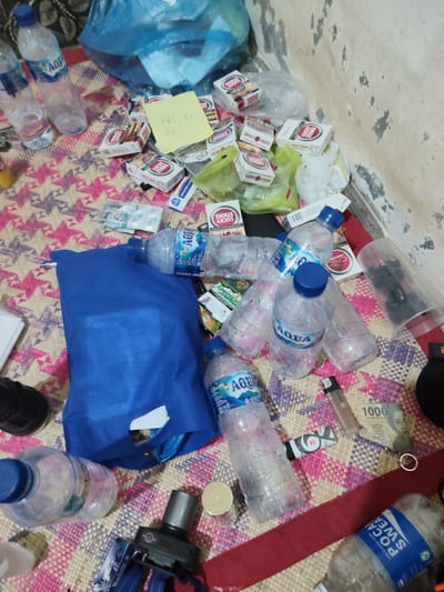 The image depicts a messy floor scene, likely indoors, with various objects scattered across a woven mat. Dominating the frame are several plastic water bottles, some appearing nearly full and others empty. Prominently visible are multiple packs of "Lucky Strike" cigarettes, arranged in piles and individually. Also present are some currency notes, a lighter, and a yellow sticky note with writing. A large blue bag is centrally located, with some items peeking out. The background shows a textured wall with discoloration. The overall impression is one of disarray and perhaps recent activity. The scene is set in the city of Mee, Indonesia.