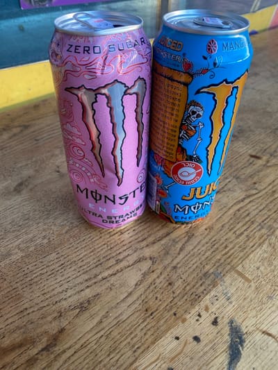 Two Monster Energy drink cans stand upright on a distressed wooden surface in Kfar Vitkin, Israel. The can on the left is pink, labeled "MONSTER ZERO SUGAR ULTRA STRAWBERRY DREAMS," with a partially lifted silver tab. The can on the right is blue, labeled "JUICED MONSTER MANGO LOCO," featuring a skeleton design and Hebrew text, also with a partially lifted silver tab. Both cans exhibit minor surface imperfections and are positioned close together, leaning slightly against each other. The wooden surface displays prominent grain patterns, scratches, and dark stains. A blurred background shows indistinct yellow and blue elements.