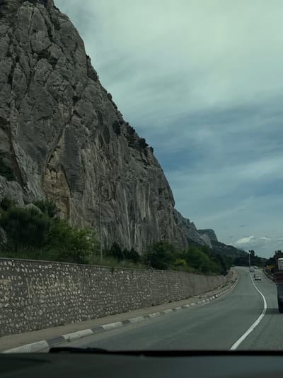 The image presents a landscape featuring a road running alongside a steep, rocky cliff face. The road is paved and marked with a white line, suggesting a two-way traffic flow. Several vehicles are visible on the road. A low, stone barrier separates the road from the cliff edge, with vegetation growing along the base of the cliff. The sky is overcast. The image is taken from a slightly elevated viewpoint, possibly from inside a vehicle. The exact location within Орлиновский округ, Ukraine, is not specified, but the scene suggests a mountainous or hilly region.