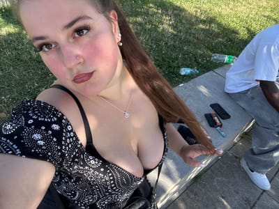 In an outdoor setting, possibly a park or public seating area in Weil am Rhein, Germany, a young woman with long brown hair is captured in a selfie. She is seated on a concrete bench, with green grass and sunlight visible in the background. She is wearing a black off-the-shoulder top with a white floral pattern and a black bra. A silver pendant necklace adorns her neck. To her right, partially visible, is another person, wearing a white t-shirt and grey jeans, seated on the same bench. The ground around the bench has some litter, including plastic bottles and cigarette butts, suggesting a public space that may not be regularly cleaned. The woman's pose and the close-up framing of her upper body indicate a casual, personal moment.