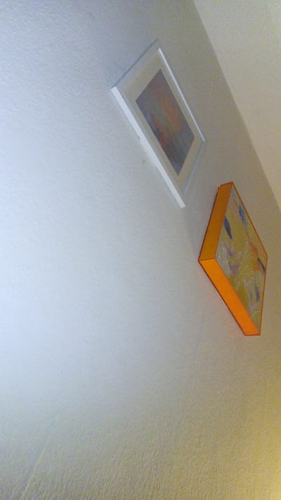 The image displays an indoor wall, likely captured from a low, upward-angled perspective, possibly highlighting a corner or the upper section of a room. The wall is painted in a light, neutral color, appearing off-white or light cream, and exhibits a subtle, textured finish across its surface.

Two distinct pieces of abstract artwork are mounted on this wall. The upper artwork is a rectangular piece enclosed in a simple, flat white frame. The artwork within the frame appears to be an abstract composition with a blend of muted colors, potentially depicting an abstract landscape or a soft play of light and shadow, though precise details are indistinct due to the distance and angle.

Below and to the right of the first piece, a second, more prominent artwork is displayed. This piece is thicker, suggesting it's a stretched canvas or a box-framed piece, notable for its bright, solid orange-colored sides. The front surface of this artwork features a vibrant abstract composition predominantly in yellow tones, accented with various abstract shapes and brushstrokes in colors such as blue, purple, and white.

The lighting in the scene indicates an indoor environment. The lower section of the wall appears to have a warmer, yellowish hue, suggesting illumination from an unseen light source or filtered natural light. There is no activity or event taking place; the scene is static. No people are visible, and there is no visible text on the artworks or the wall. Although the provided location context is Ginsheim-Gustavsburg, Germany, the image itself offers no specific architectural or environmental cues to identify this particular location, presenting a generic interior setting.