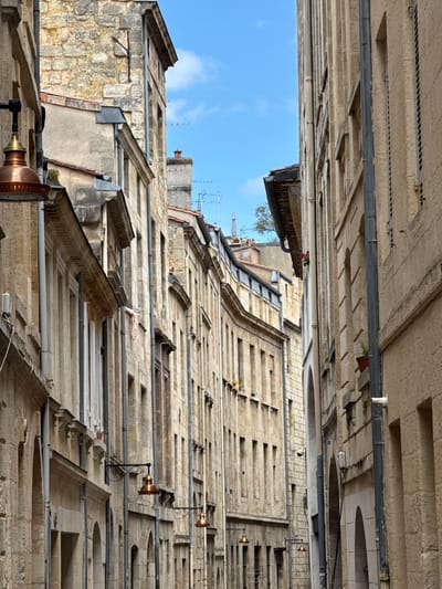 The image depicts a narrow street in Bordeaux, France, with tall, aged stone buildings lining both sides. The architecture suggests a historical European city, characterized by its weathered facades, numerous windows, and intricate stonework. Several gas-style street lamps with copper-colored shades are visible, attached to the buildings, hinting at a blend of old and modern infrastructure. The street is largely empty, contributing to a sense of quietude. Above, a sliver of bright blue sky with scattered clouds is visible, indicating daylight. The scene captures the atmospheric charm of Bordeaux's historic districts.
