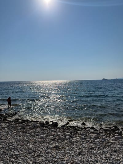 A person stands at the edge of a rocky shoreline, with the calm sea stretching out before them. The sun, visible in the upper left of the frame, casts a bright glare on the water, creating a shimmering effect. The sky is clear and blue, suggesting fair weather. The scene appears to be a tranquil beach, with the sun's rays indicating it's likely daytime, possibly late afternoon given the angle of the light. The water is a deep blue, with small waves gently lapping at the shore. The shoreline itself is composed of numerous gray and brown pebbles, varying in size. The person is silhouetted against the bright water, making their features indistinguishable. They appear to be looking out at the sea, perhaps contemplating the view or enjoying the peaceful atmosphere. There is no visible text in the image.