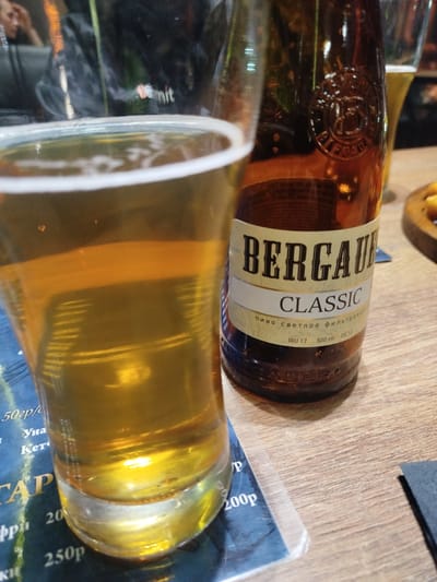 The image shows a close-up of a glass of beer and a bottle of Bergaur Classic beer on a wooden table. The beer is a light golden color, and the glass is filled nearly to the top. The bottle of Bergaur Classic beer has a label with the words "Bergaur" and "Classic" clearly visible. The table is wooden. The photo was taken indoors, likely in a bar or restaurant. There is a menu visible with prices in Russian rubles. Gus-Khrustalny is the location.