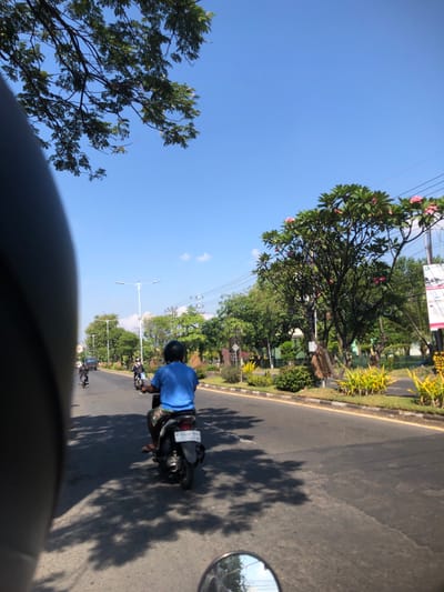 The image depicts a street scene in Buduran, Indonesia, under a clear blue sky. A person wearing a blue shirt and a black helmet rides a motorcycle down the asphalt road. The motorcycle has a white license plate with black lettering. The road is lined with green trees and decorative bushes with yellow and pink flowers. Streetlights are visible on the side of the road. The perspective suggests the photo is taken from another motorcycle, with a portion of a black helmet visible in the foreground. Shadows of trees are cast across the road. Other motorcycles are also seen in the distance.