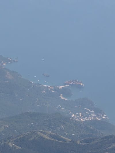 The image is an aerial view of a coastal area with mountains and the sea. In the foreground, lush green mountains slope down towards the coastline. Scattered on the mountainside and along the shore are several buildings, appearing to be a resort or a developed area with a cluster of white and light-colored structures.
The coastline is irregular, with several inlets and rocky outcrops. The sea is a calm, pale blue, with several small white objects that could be boats or buoys visible on the surface. Further out at sea, there is a small island or a very large rock formation, also appearing to be developed with buildings. A curved sandy beach is visible in one of the inlets. The overall impression is one of a scenic, developed coastal landscape.
The scene is likely located in the vicinity of Donja Cijevna, Montenegro, given the mountainous coastal terrain and the architectural style of the buildings which can be found in such regions. The time of day appears to be daytime, with clear, possibly hazy conditions contributing to the soft lighting. There are no people visible in the image.
