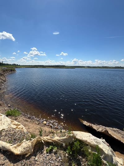 The image depicts a waterfront scene in Yoksolovo, Russia, under a clear blue sky with scattered white clouds. The foreground shows a rocky shoreline with some scattered debris, including what appears to be a large piece of torn, beige fabric or tarp. Further down the shore, there is a pile of rocks and some green vegetation. The dark blue water of a lake or river occupies the majority of the frame, with gentle ripples reflecting the sunlight. In the distance, a line of trees forms the horizon, with more trees and greenery visible on the far bank. The overall environment suggests an outdoor, natural setting. There are no people or animals visible in the image. The context of the location being in Yoksolovo, Russia, provides a geographical identifier for the scene.
