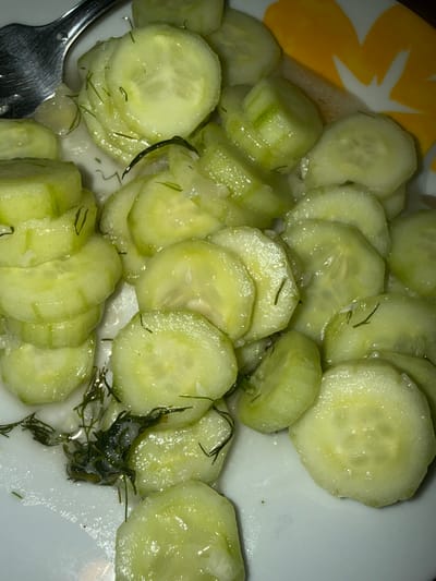 A close-up, overhead shot captures a plate of thinly sliced cucumbers dressed with a creamy sauce and dill. The cucumbers are a pale green, some showing slight translucency from being sliced. Small dark green flecks of dill are scattered throughout the dish, and glistening droplets of sauce coat the vegetables. In the lower left corner, a silver fork is partially visible, its tines resting on the edge of the plate. The plate itself is white and appears to be slightly chipped on the left side, revealing a bit of a yellow and white floral pattern beneath. The background is a blurry mix of white and yellow, suggesting the plate is part of a larger placemat or serving dish. The lighting is overhead and somewhat dim, creating subtle highlights on the moist cucumber slices. There is no discernible text or specific time of day, weather, or emotional cues. The setting appears to be a casual meal.