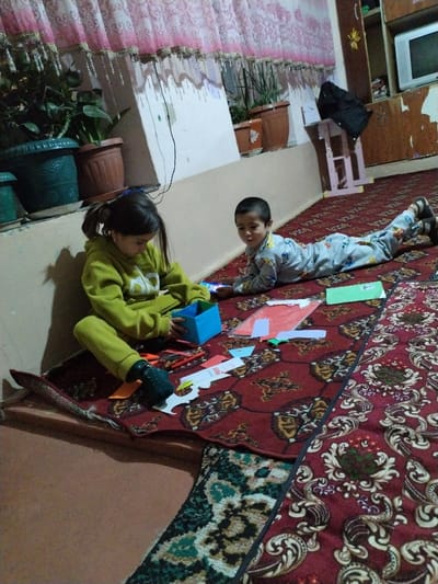 The scene depicts an indoor residential setting, likely in Limbur, Uzbekistan. Two children are present on a floor covered primarily by a large, ornate red rug with white and gold patterns.

In the foreground, a female child, approximately 6-8 years old, is seated cross-legged on a smaller patterned rug, which is placed over a plain brown floor section and partially beneath the main red rug. She wears a lime-green hooded sweatshirt and matching pants, with dark socks. Her hair is styled in two ponytails. She is actively engaged in a craft activity, holding a small blue cardboard box with both hands. Various craft supplies are scattered around her on the rug, including orange scissors, red and green markers, several pieces of colored paper (red, green, blue, orange), and white paper cutouts, one resembling a stylized animal or figure.

To the right and slightly behind her, a male child, approximately 2-4 years old, is lying prone on the red rug, facing towards the viewer. He wears light-colored pajamas with multiple cartoon character patterns and dark shoes or slippers. His head is slightly lifted, and he appears to be observing the scene or the camera.

The background features a light-colored wall with a lower dark baseboard. Above the baseboard, several potted plants are arranged, including a large plant in a dark green pot and smaller plants in terracotta-colored pots. Pink and white patterned curtains with decorative tassels hang above the plants, extending across the upper portion of the wall. To the far right, a light-colored wooden stool with a black garment draped over it is visible. Above the stool is a multi-tiered wooden shelving unit or cabinet, which appears somewhat worn, with peeling veneer. A small, older model television set with a grey screen is situated within one of the shelves. A white electrical cord hangs down the wall near the plants.