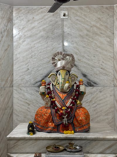 The image depicts a statue of the Hindu deity Ganesha, situated on a marble platform. The statue is adorned with an elaborate headdress, floral garlands, and an orange and blue robe. The platform rests against a wall covered in white marble. A light switch is visible on the wall to the left of the statue. A small offering plate and a box are positioned on the platform next to the statue. This scene is likely within a temple or shrine. While the statue resembles Ganesha, the lack of further identifying information makes it difficult to confirm the location in Vadodara, India.