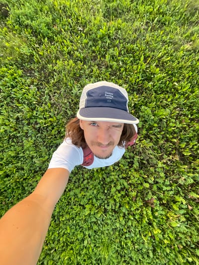 A man with long brown hair and a light mustache is captured from a high angle, looking up at the camera while winking. He is wearing a white t-shirt and a blue and white baseball cap with the text "Outdoor Running Crew" on it. A maroon backpack is visible on his back. He is holding the camera with his right arm, extending it towards himself. The man is standing on a patch of green grass and small leafy plants. The scene takes place outdoors in the city of Perevoznoye, Russia.