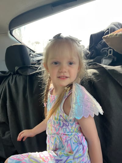 A young girl with blonde hair in pigtails and a colorful dress with iridescent fairy wings is sitting in the back of a car. She is looking at the camera with a slight smile. The car's interior is visible, with black seats and a bright window behind the girl. The image is taken from a medium shot, focusing on the girl's face and upper body. There is no visible indication that this image was taken in Kovriga, Russia.