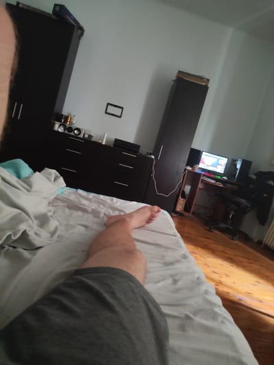 The image depicts a bedroom scene from a first-person perspective, likely taken with a smartphone. The viewpoint is from a bed, looking towards a dresser, a wardrobe, and a desk with a computer setup. A person's legs and torso, dressed in grey shorts, are visible in the foreground, resting on white and grey bedding. The room has light blue walls and a wooden floor.
On top of the dark brown dresser, several items are present, including what appear to be speakers, bottles, and containers. To the left, a tall, dark brown wardrobe with double doors is visible. To the right, a wooden desk holds a computer monitor displaying a colorful image, a desktop tower, a keyboard, and a mouse. A black office chair is positioned at the desk. A tall, dark brown cabinet or wardrobe is situated behind the desk. The lighting suggests it is daytime, with sunlight streaming in from the right, casting shadows on the floor.
The image does not contain any specific visual cues that would allow for definitive identification of the location within the city of Arad, Romania.