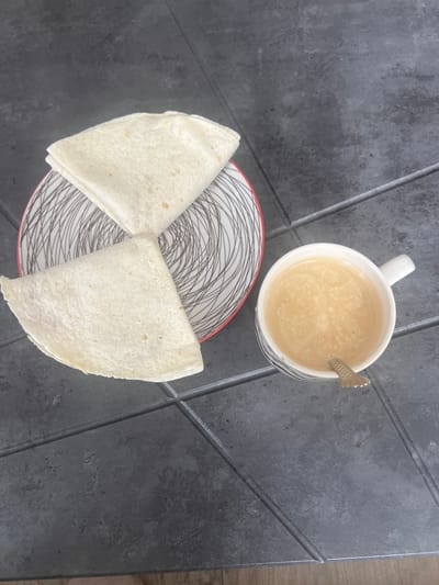 A top-down, eye-level shot captures a simple breakfast setup on a dark, textured surface, possibly a table with tiled sections. In the upper right portion of the frame, a white ceramic mug filled with a light brown, frothy beverage, likely coffee or a latte, rests. A small, metallic spoon is partially submerged in the drink. Below the mug, a round plate with a black and white geometric pattern is visible. Two white tortillas, folded in half, are arranged on the plate to form a loose bow shape. The tortillas appear soft and slightly puffed, with a few small, brown speckles on their surface. The overall scene suggests a casual meal, possibly breakfast or a mid-day snack. The lighting appears to be natural, casting soft shadows, and the setting does not provide specific cues about the time of day beyond suggesting it is daytime. There is no visible text in the image. The context indicates the location is Magnitogorsk, Russia.