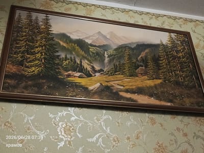 A framed oil painting of a mountainous landscape with a waterfall and two small houses is displayed on a wall with floral wallpaper. The painting depicts a serene scene with tall evergreen trees, rolling hills, and majestic mountains in the background. A dirt path winds through the landscape, leading to the houses. The wallpaper is a pale green with a repeating pattern of beige and white flowers and swirling vines. In the bottom left corner of the image, a timestamp reads "2026/06/28 07:39" and the word "храмов" is written below it. The location is specified as the city of Yalta, Ukraine.