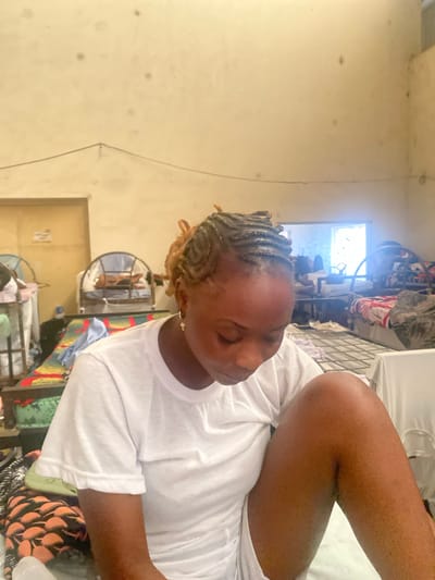 A young Black woman with braided hair and a white t-shirt is seated in a room. She is looking down and has her right leg bent up towards her chest. The room appears to be a dormitory or shared living space, with multiple beds visible in the background. The beds are made with colorful linens and some have mosquito nets. The walls are a pale yellow and show signs of wear and tear. There is a window in the background that lets in natural light. The overall impression is one of a simple and functional living environment. The image was taken in Jimeta, Nigeria.