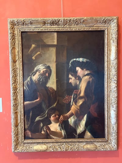 This is a horizontal, framed oil painting depicting a scene with several figures. The painting is oriented such that the bottom edge of the frame is to the left and the top edge is to the right. The style appears to be Baroque, with dramatic lighting and detailed rendering of human forms and drapery.
In the foreground, an older man with long, graying hair and a beard is visible, his head turned to his right. He wears dark clothing and his face is lit from above, creating shadows that emphasize his features. His expression is somber. To his left, two cylindrical objects, possibly goblets or small vases, stand on a surface.
Behind him, in the middle ground and upper portion of the painting, two other figures are depicted. One figure, partially obscured, appears to be leaning over and touching something. Another figure, with fair skin and dark hair, is looking downwards, with their hands engaged in an action. They are dressed in light-colored fabric, possibly a shift or tunic, and a striped head covering. The interaction between these figures is unclear but suggests a moment of shared activity or attention. The background is dark and indistinct, providing little information about the setting. The overall mood is serious and contemplative.
The painting is enclosed in an ornate, gilded frame with decorative carvings, featuring floral motifs and recessed panels. The background wall on which the painting is displayed is a vibrant coral or salmon color. No visible text is present on the painting or frame. The image itself has a slight tilt, consistent with a photograph taken of a painting hung on a wall.