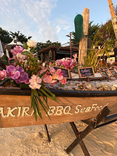 An outdoor seafood display is positioned on a sandy beach in Ban Ko Li Pe, Thailand, illuminated by warm late afternoon or early evening sunlight. The primary subject is a weathered wooden boat, serving as a display stand, supported by dark metal cross-stands. The boat's side features carved and white-painted text reading "AKIRA BBQ SeaFood."
Within the boat, various seafood items are arrayed on a bed of ice. Small, freestanding chalkboards indicate offerings and prices, including "Oysters 3 pcs / 550.-," "Jackfish 800," and "Red Snapper 600 kg." The boat is decoratively adorned with a large arrangement of artificial flowers, primarily pink and purple orchids, interspersed with white roses and green foliage.
In the mid-ground, behind the seafood display, several structures characteristic of a beachside resort or restaurant are visible. To the left, a structure features a white conical roof. Centrally, a large, rough-hewn wooden post stands, draped with a dark green fishing net-like material, with a light fixture attached to an adjacent pole. Further back, a rustic wooden building with a thatched or wooden roof is present. Tropical vegetation, including palm trees, is distributed throughout the background. A single, blurry individual is discernible seated at a table under a thatched roof structure in the far right background. The sky overhead is clear blue with scattered white clouds.