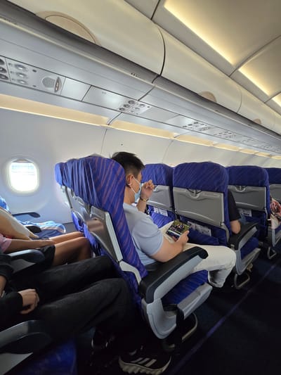 The image captures the interior of an airplane cabin, specifically focusing on the seating area. The seats are upholstered in blue fabric with a patterned design and have gray headrests and armrests. Several passengers are visible, including a man in the foreground wearing a light blue shirt and a face mask, who is holding a tablet. Other passengers are seated behind him, with some partially obscured. The overhead compartments and lighting system of the airplane are also visible. The overall environment suggests a typical commercial flight. The image does not provide any information that would allow for determining its location within Xiaoshan District, China, or any other city.