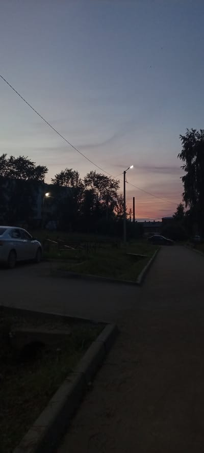 This vertical, low-angle, outdoor, dusk shot captures a street scene in Kudymkar, Russia. The sky is a gradient of pale blue to soft pink and orange, with a few faint clouds. A dark power line stretches diagonally across the upper portion of the frame. In the mid-ground, silhouetted trees line the horizon, obscuring a building behind them. A solitary street lamp illuminates the scene, casting a glow on the pavement and surrounding area. To the left, the rear of a light-colored sedan is visible parked on a paved area. Further to the right, the road curves, with another dark-colored car parked in the distance. The foreground features a paved road with a concrete curb and grass. The overall impression is a quiet, transitional moment at the end of the day.
