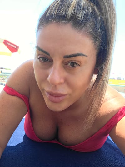 The image is a close-up, selfie-style photograph of a woman with her head tilted downwards, looking towards the camera. She has long, blonde-streaked hair pulled back into a high ponytail and is wearing a pink off-the-shoulder top or bikini. Her skin appears sun-kissed and has visible freckles. The background suggests an outdoor setting, possibly a beach or poolside, with a bright sky and what appears to be a yellow and red umbrella or awning on the left side of the frame. The overall lighting is bright, indicating it was taken in daylight. There is no discernible connection to the city of Reggio nell'Emilia, Italy, in the image itself.
