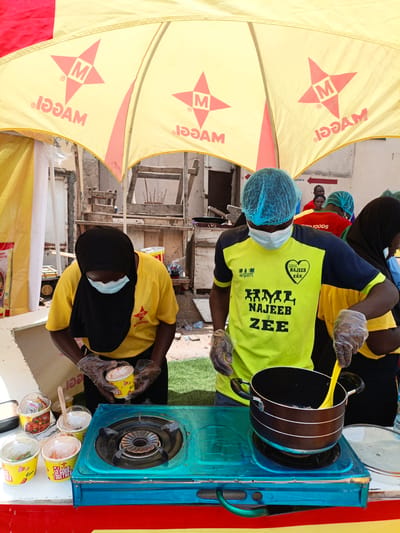 This image captures a street food stall in Tarauni, Nigeria, under a bright yellow umbrella advertising "Maggi." Two individuals, dressed in bright yellow t-shirts and wearing hairnets and face masks, are actively preparing food. One person, wearing a black hijab and a yellow t-shirt, is holding a cup of what appears to be a food product, possibly noodles or soup, while the other, in a neon yellow and navy t-shirt, uses a bright yellow spoon to stir something in a black pot on a teal gas stove.

Several small cups with clear lids are lined up on the counter, some containing food items like vegetables and what looks like a red curry. The text on the cups includes "SO GOOD TOGETHER" and "Maggi." The background shows a somewhat rustic setting with wooden structures and glimpses of other people, some also wearing hairnets, suggesting a communal or event-based food service. The sunlight indicates it's daytime, and the general atmosphere appears busy and focused on food preparation and service. The "Maggi" branding is prominent on the umbrella, suggesting this is a promotional event or a vendor associated with the brand.