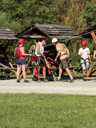 Four individuals, three males and one female, are gathered outdoors in front of wooden structures. The female, on the far left, wears a red helmet, a red backpack, and blue shorts. She holds a red helmet in her right hand. To her right, a male in a white sleeveless shirt and black shorts is adjusting his gear, holding a red helmet. Further to the right, another male in a tan t-shirt, white helmet, and olive-green shorts is also attending to his equipment. The fourth individual, a male on the far right, wears a white t-shirt, light blue shorts, and a red helmet. All four appear to be wearing harnesses and safety gear, suggesting an activity like zip-lining or climbing. The background consists of dense green foliage and trees. The ground is covered in gravel. The overall setting suggests an outdoor adventure or recreational facility. The location of Warisy, Belgium is not discernible from the visual information in the image.
