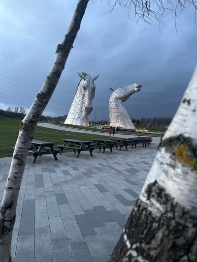The image depicts The Kelpies, two colossal horse head sculptures, located in Falkirk, United Kingdom. The sculptures, constructed from interwoven stainless steel plates, dominate the mid-ground against an overcast, grey sky. The left sculpture faces forward, while the right one faces slightly right and upwards. Both are illuminated from below.
In the foreground, a paved plaza composed of alternating light and dark grey rectangular pavers extends from left to right. A row of at least eight dark wooden picnic tables with attached benches is arranged linearly on this paved area, stretching into the mid-ground.
Two birch tree trunks partially frame the scene; one on the left foreground runs vertically, obscuring a portion of the view, and another on the right foreground is partially visible. Beyond the paved area and picnic tables, a manicured green grass slope rises gently towards the sculptures.
Several individuals are visible in the mid-ground, near the base of The Kelpies. Two figures, possibly adults, are walking between the sculptures, and another figure is visible further right, near the end of the picnic table row. Distant bare trees line the horizon on the left and right, with power lines and a pylon visible in the far left background. The overall lighting suggests dusk or a heavily overcast day.
