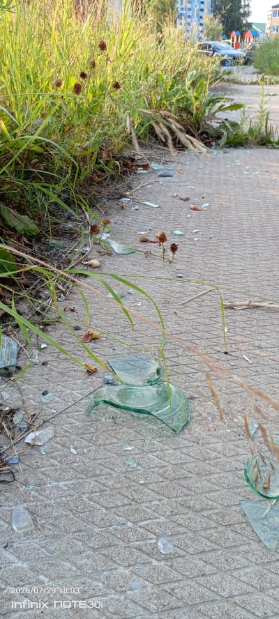 The image depicts a section of an outdoor concrete walkway, possibly a sidewalk or path, in the city of Langepas, Russia. The walkway is textured with a diamond pattern. Along the edge of the walkway, tall green grass and dry weeds are growing, encroaching onto the concrete surface. Scattered across the concrete are numerous shards of broken green glass, indicating potential vandalism or an accident involving a glass object. In the background, out of focus, are elements of an urban environment, including a dark-colored car, a playground with colorful structures, and buildings. The scene appears to be captured during daylight, with sunlight illuminating the vegetation and casting shadows. The bottom of the image displays a timestamp and phone model: "2026/07/29 18:03 Infinix NOTE 30i".
