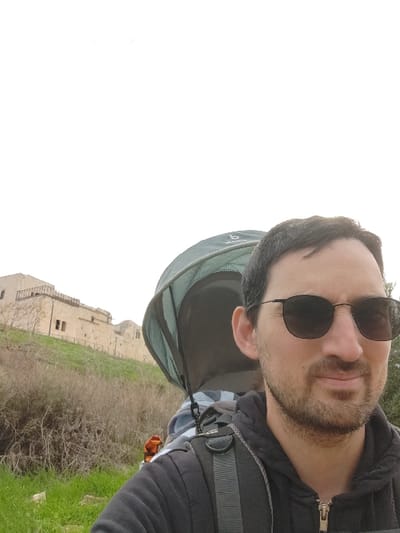 The image depicts an outdoor, slightly upward-angled selfie-style photograph taken near Antipatris Fortress in Rosh HaAyin, Israel. In the right half of the frame, a male subject in his 30s-40s is visible, wearing dark sunglasses, a short beard, and a black zippered hoodie. He is carrying a dark green child-carrying backpack with a deployed sun canopy on his back; light-colored patterned fabric is visible inside the carrier, indicating it is in use, though a child is not explicitly visible. A small orange object is attached to the left side of the carrier. The foreground shows green grass on the lower left, transitioning to dry, sparse brown vegetation covering a hillside. In the mid-left background, on an elevated position, stands a large, light-colored stone structure identified as Antipatris Fortress (also known as Binar Bashi or Ras al-Ayn Fortress), featuring multiple windows, arches, and a fortified upper section. The sky overhead is uniformly bright white and overcast.