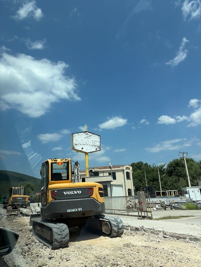 A yellow Volvo excavator, model GENEX 65, is parked on a gravel lot next to a building. The excavator has a black undercarriage and tracks, and a black Volvo logo is visible on its side. A white hexagonal sign with the text "CARINVEST" is mounted on a yellow pole behind the excavator.

The scene appears to be a construction or industrial area. In the background, there are more excavators, piles of materials, trees, and a building with white siding. A power line pole is also visible. The sky is blue with scattered white clouds. The image seems to be taken on a sunny day.

The orientation of the image is rotated 90 degrees clockwise. The location is Lastva Grbaljska, Montenegro. No people are visible in the image.