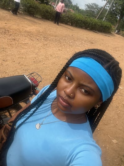 An adult female with dark skin, black braided hair, a light blue fabric headband, and a light blue short-sleeved top is depicted in a close-up, upward-angled shot. She wears a small gold-colored stud piercing in her right nostril, small gold-colored hoop earrings, and a silver-colored chain necklace with a Hello Kitty pendant. To her left, the rear section of a black motorcycle, including its seat and a chrome grab handle, is visible. The immediate ground is unpaved, consisting of light brown earth mixed with small stones. In the mid-to-background, two adult males are present in the city of Osu, Nigeria. One male, partially obscured on the left, wears a white top and dark trousers, appearing to walk left. Another male, further right, wears a light pink long-sleeved top and dark trousers, standing and facing left. A low line of green foliage separates the immediate unpaved area from a distant paved surface, utility poles, and mature trees visible under a bright sky.
