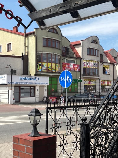 This is a vertical, eye-level photograph of a street scene in Mińsk Mazowiecki, Poland. The image is dominated by the facades of several buildings lining the street, with a wrought-iron gate in the foreground partially obscuring the view. The buildings are of varying architectural styles, with some featuring modern renovations and others older, more traditional designs. Red-tiled roofs are visible on several structures, and the buildings have multiple stories with numerous windows.
The street itself appears to be paved, with visible lane markings. A dark-colored car is parked on the right side of the street, and a bicycle is leaning against a storefront. A pedestrian crossing sign, featuring a silhouette of an adult and child walking and a bicycle symbol, is prominently displayed on a blue circular sign mounted on a pole.
Several signs with text are visible on the buildings. One large white sign reads "Este Medicus KLINIKA MEDYCYNY ESTETYCZNEJ - CENTRUM MEDYCZNE," indicating a medical or aesthetic clinic. Other signs advertise clothing ("Tania Odzież," "Tania") and mention sales ("50%"). Another sign features the word "CHINY" in large red letters.
The sky above is mostly clear, with some wispy clouds, suggesting it is daytime. The lighting suggests it is likely midday or early afternoon. The overall impression is of a moderately busy urban street with various commercial establishments.