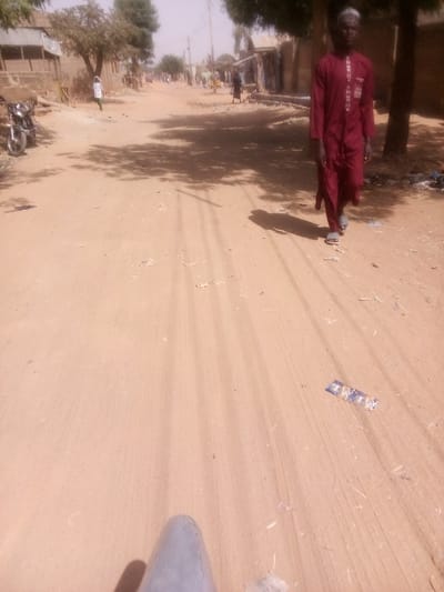 The scene depicts an unpaved, light brown dirt road in Rangaza, Nigeria, under bright, possibly hazy, daylight. In the immediate foreground, a partial view of a grey, conical object, consistent with a vehicle component, is visible. Distinct tire tracks traverse the dusty road surface. A man, wearing a dark red embroidered tunic (kaftan), a white cap, and light-colored sandals, walks from the right side of the frame towards the left, casting a shadow directly behind him. Approximately ten feet in front of the man, a blue and white wrapper lies flat on the road.
To the left of the road, a silver motorcycle is parked. Behind it, a series of single-story structures with mud-colored walls and corrugated metal roofs are visible. Scattered green trees and their shadows are present on both sides of the road. Further down the road on the left, another individual in light-colored clothing is walking. The right side of the road also features additional low-rise buildings and trees. The sky is a pale, hazy blue. No direct interactions between individuals are discernible.