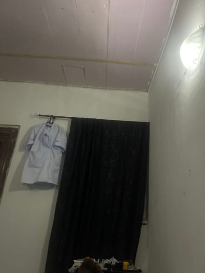 The image depicts an interior scene, likely a bedroom or living space in Tavo, Nigeria. A white overhead light fixture is visible on the ceiling, emitting a bright glow. The walls appear to be a light color, with one section on the left covered in large, horizontally oriented panels that are a light pink or purple hue. These panels are joined by a vertical strip of tape.

In the center-right of the image, a black curtain is drawn across a window or wall. Hung on a rack beneath the curtain is a white shirt, possibly a uniform or formal wear. The shirt is folded or draped in a way that suggests it is either new, has just been ironed, or is being aired out.

The overall lighting is somewhat dim, with the ceiling light being the primary source. The angle of the photo is slightly tilted, giving a dynamic perspective of the room. There are no visible people, but the presence of the shirt suggests a recent or imminent human activity. The scene is calm and appears to be indoors. No text is visible in the image.