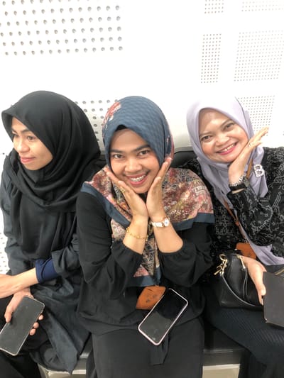 Three women wearing hijabs are sitting on a black bench. The woman on the left is wearing a black hijab and a black outfit, looking to her left. The woman in the middle is wearing a patterned blue and brown hijab and a black top, smiling at the camera with her hands on her cheeks and braces visible. The woman on the right is wearing a light purple hijab and a black patterned top, smiling at the camera with her hand near her face. All three women are holding smartphones. A black purse is resting on the lap of the woman on the right. The background consists of a white wall with a perforated pattern. The scene appears to be indoors, possibly in a waiting area or public space. Based on contextual information from Reuleut Timu, Indonesia, this location could be an administrative building, a community center, or a transport hub.