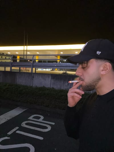 A man wearing a black "47" cap and black shirt is seen smoking a cigarette. The image is taken at night, with a lit building visible in the background, reflecting on the metal railing in front of it. A "STOP" sign is painted on the dark ground, next to a hedge, adding context to the location. The photograph is shot at an angle, placing the subject on the upper portion.