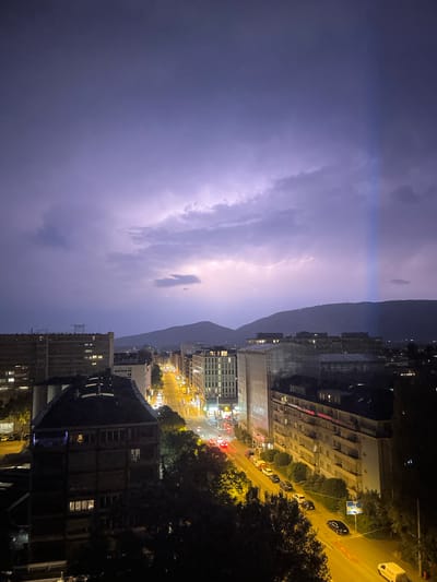 The image depicts a nighttime urban scene in Geneva, Switzerland, with a dramatic thunderstorm occurring. The perspective is from a high vantage point, looking down and across a city illuminated by streetlights and building lights, with a mountain range visible in the background.

The primary subject is the powerful lightning striking through the dark, cloudy sky. Streaks of brilliant white light illuminate the clouds and cast a purplish glow over the city. This storm creates an atmosphere of awe and intensity.

Below the stormy sky, the city unfolds. Buildings of various sizes and architectural styles are visible, many with windows lit, suggesting human activity within despite the late hour and inclement weather. A street runs through the scene, lined with parked cars and trees. The streetlights cast a warm, yellow hue, contrasting with the cool, electric blue of the lightning. The presence of cars, streetlights, and illuminated buildings firmly places the scene in an urban environment.

The time of day is clearly night, emphasized by the darkness, the artificial lights, and the thunderstorm. The weather is a significant element, with the heavy clouds and dramatic lightning being the focal point of the sky.

Notable details include the scale of the buildings and the vastness of the storm. The mountain range in the background provides a sense of scale and potentially a geographical marker for Geneva. The contrast between the natural spectacle of the storm and the man-made city below is striking. There is no visible text in the image. The overall mood evoked is one of powerful natural forces interacting with a human settlement.