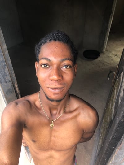 A dark-complexioned male individual is depicted in a frontal medium shot, standing shirtless within an indoor setting in Oraukwu, Nigeria. He wears a gold-colored chain with a detailed cross pendant around his neck. His short, dark hair is styled in small braids or twists, and he has light stubble on his chin, looking directly at the camera with a slight smile. The background features a dimly lit, unadorned interior with exposed concrete walls. A dark door frame or wall segment is visible on the right, featuring a silver-colored C-shaped handle. On the floor in the mid-background, to the right of the individual's head, a black, round object resembling a bowl or pan is present.