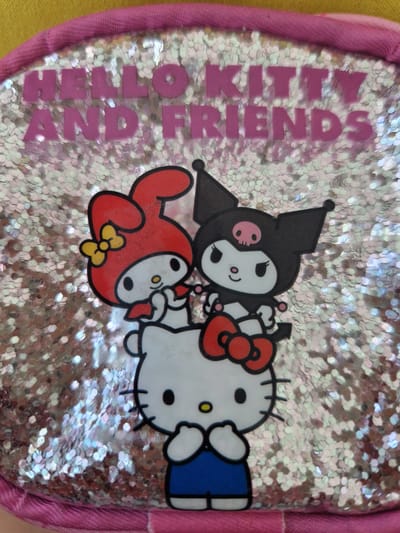 The image displays a close-up of a pink and yellow purse featuring a glittery, iridescent background. Prominently displayed on the purse are three Sanrio characters: Hello Kitty in the front, wearing a blue dress, and My Melody and Kuromi layered behind and slightly above her. The text "HELLO KITTY AND FRIENDS" is printed in pink lettering across the top of the purse. There is no information about the location in Fresnes, France within the image.